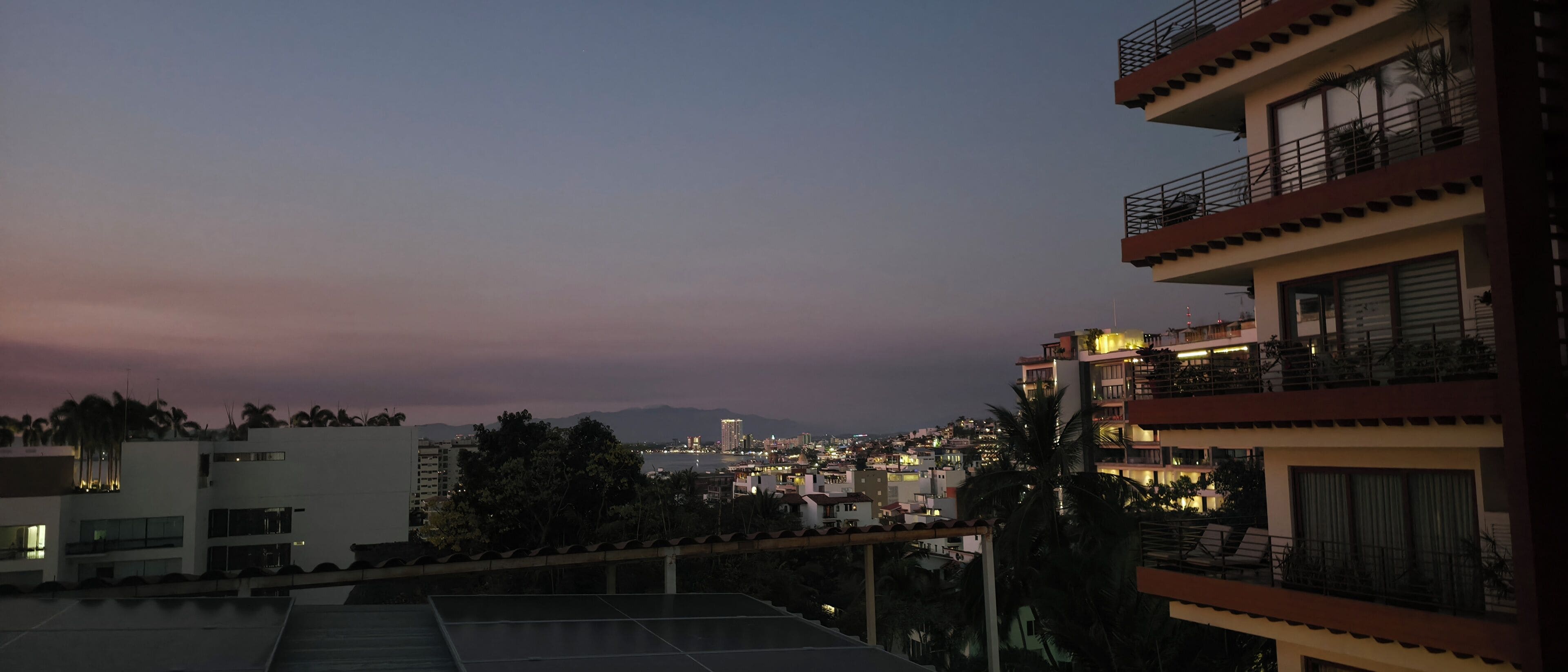 Un edificio de varios pisos con balcones, un espacio en la azotea y vista a un paisaje urbano al atardecer.