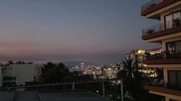 Un edificio de varios pisos con balcones, un espacio en la azotea y vista a un paisaje urbano al atardecer.