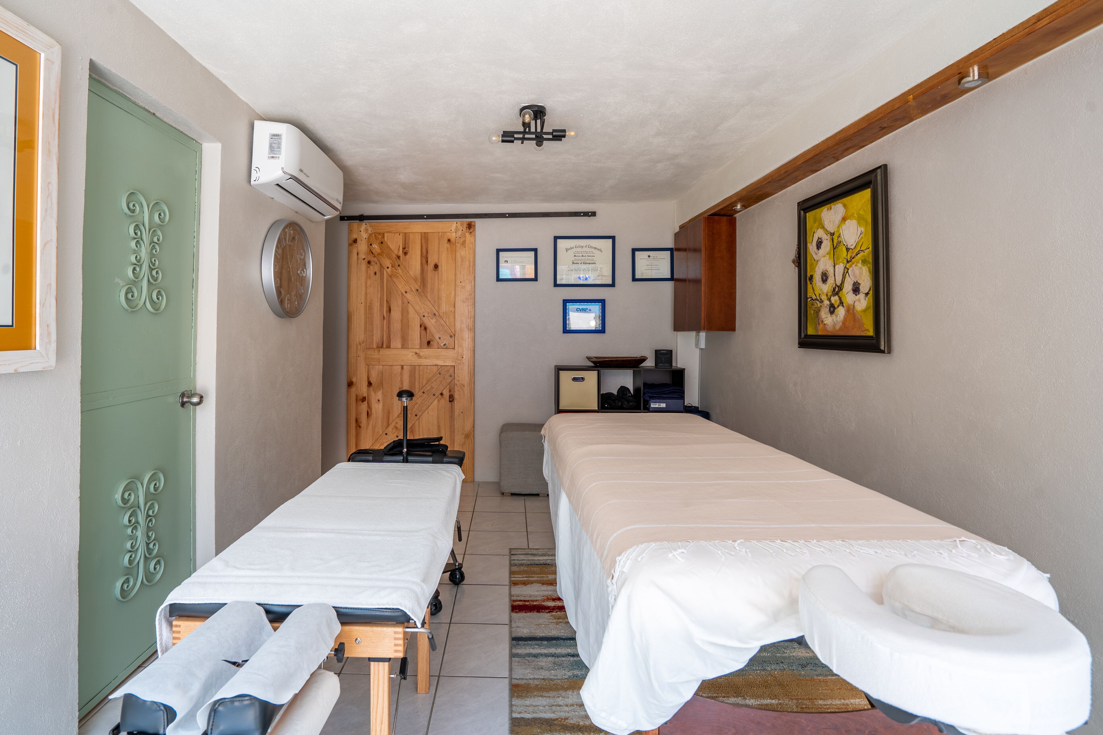 A room with two beds, a wooden door, an air conditioner, framed certificates, and a painting on the wall.