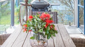 Una mesa de madera con un florero de vidrio con flores rojas en un balcón que ofrece vistas a una parrilla y follaje.