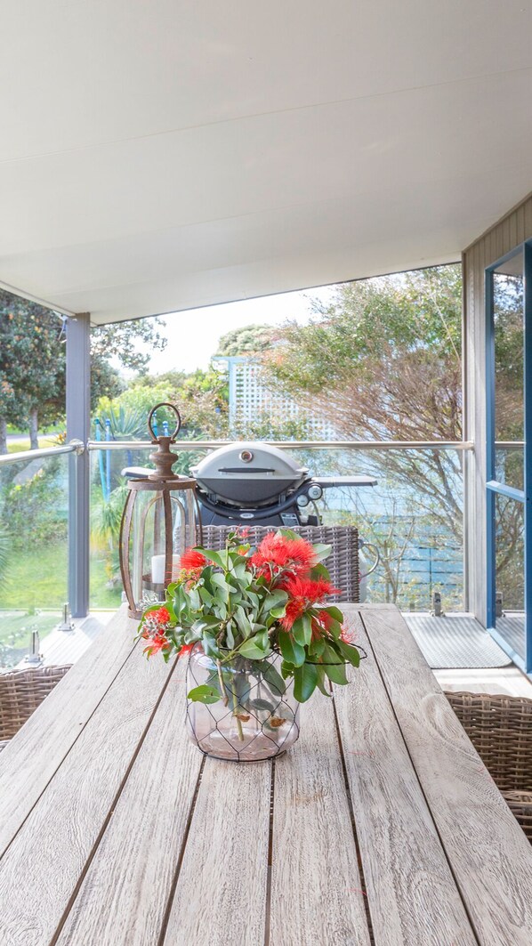 A wooden table with a glass vase of red flowers on a balcony with a view of a barbecue grill and greenery.