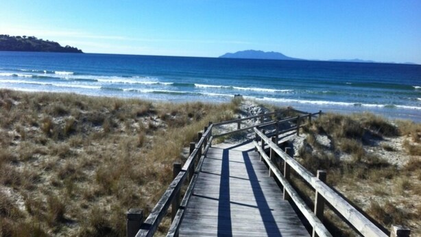 Pasarela de madera que conduce a la playa, con vista despejada al océano y montañas distantes.