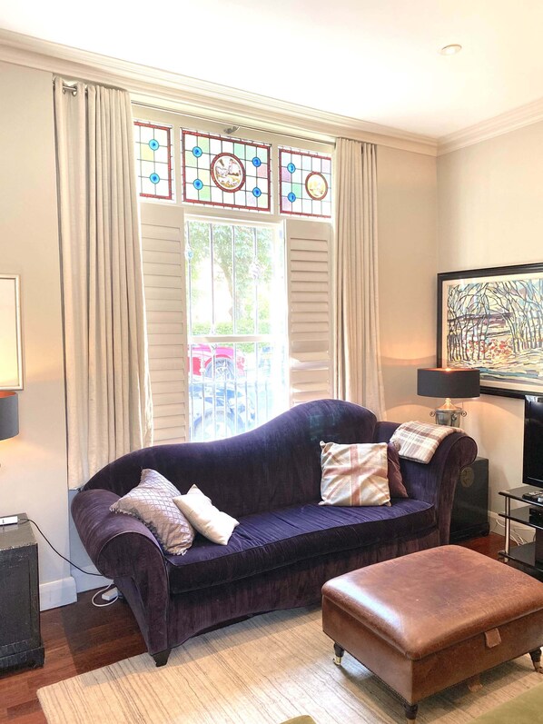A living room with a purple sofa, an ottoman, a TV, and a painting on the wall.