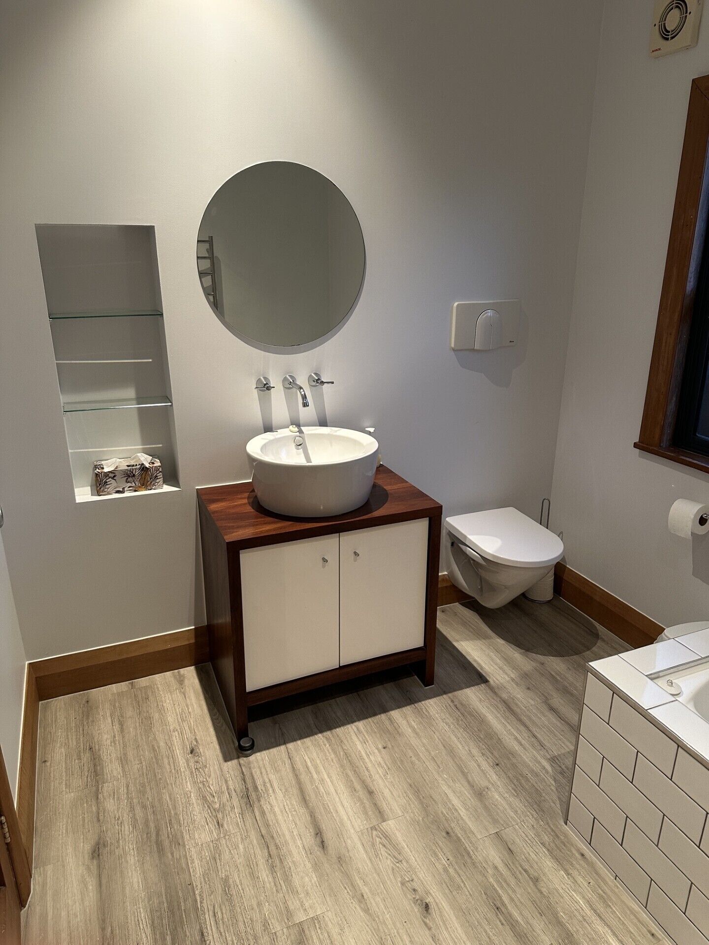 Une salle de bain avec un miroir rond, un lavabo blanc, des toilettes et une petite étagère sur laquelle se trouve une boîte de mouchoirs.