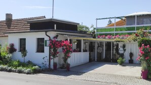 Ein weißes Gebäude mit einem Ziegeldach, einem überdachten Eingang und Üppig bepflanzten Kübeln mit rosa Blüten.