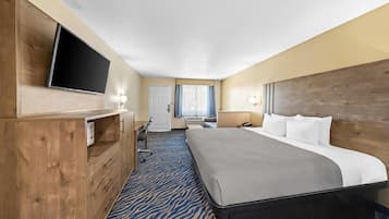 A hotel room with a large bed, wooden headboard, a flat-screen TV mounted on the wall, a desk with a chair, and a window with curtains.