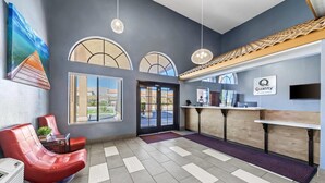 A hotel reception area with a modern design, including a reception desk, a seating area with red chairs, and large windows providing views of the outdoors. - Quality Inn & Suites Victorville – Hesperia (Hesperia)