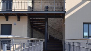 Une passerelle en bois mène à un bâtiment doté d’un escalier en pierre et d’une rambarde en métal.