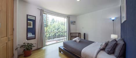 A bedroom with a large window, a bed, a bedside table, a lamp, and a potted plant.