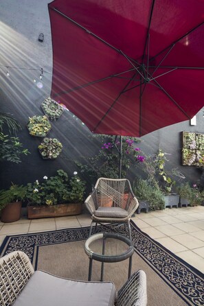 Eine Terrasse mit rotem Sonnenschirm, Korbmöbeln, Blumentöpfen und einer Zierwand.