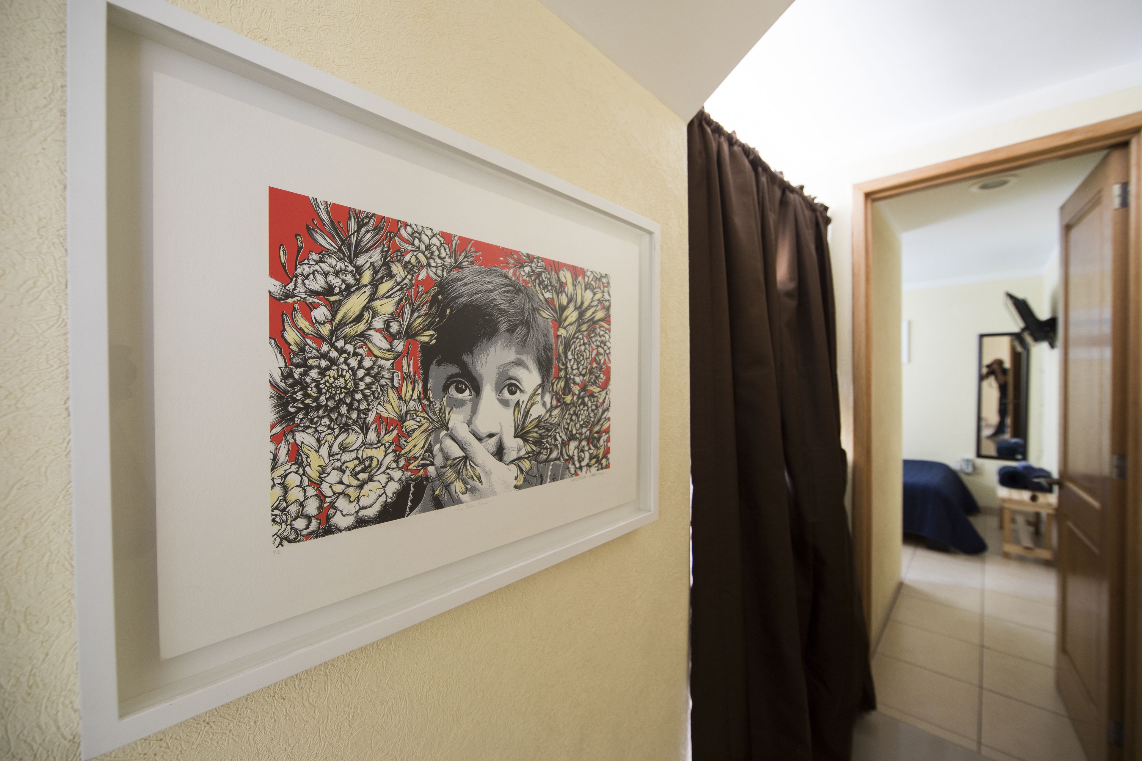 Een lijstwerk aan de muur, een deur die leidt naar een kamer met een bed en nachtkastje, en een bruin gordijn.