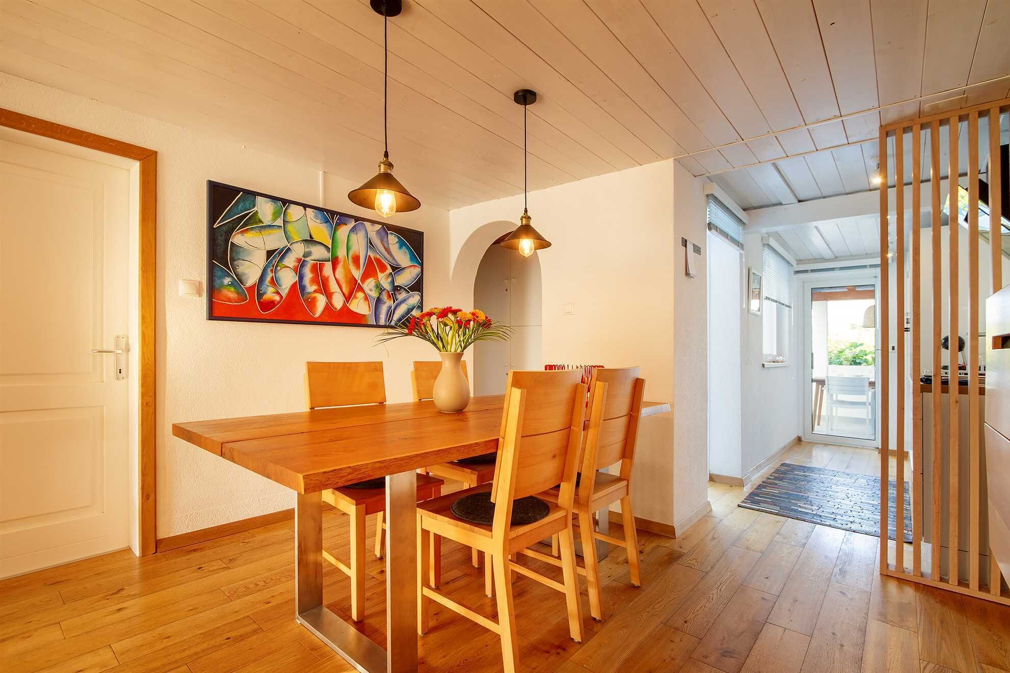 Een eetruimte met een houten tafel, stoelen, hanglampen en een kleurrijk schilderij aan de muur.