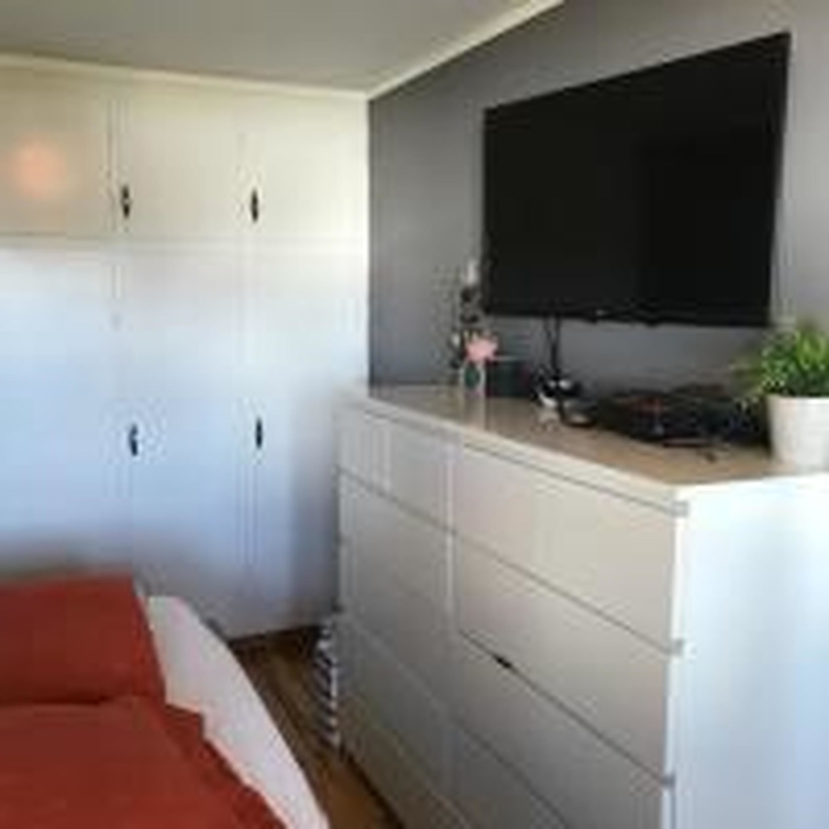 A bedroom with a bed, a dresser, a television on the wall, and a small plant.