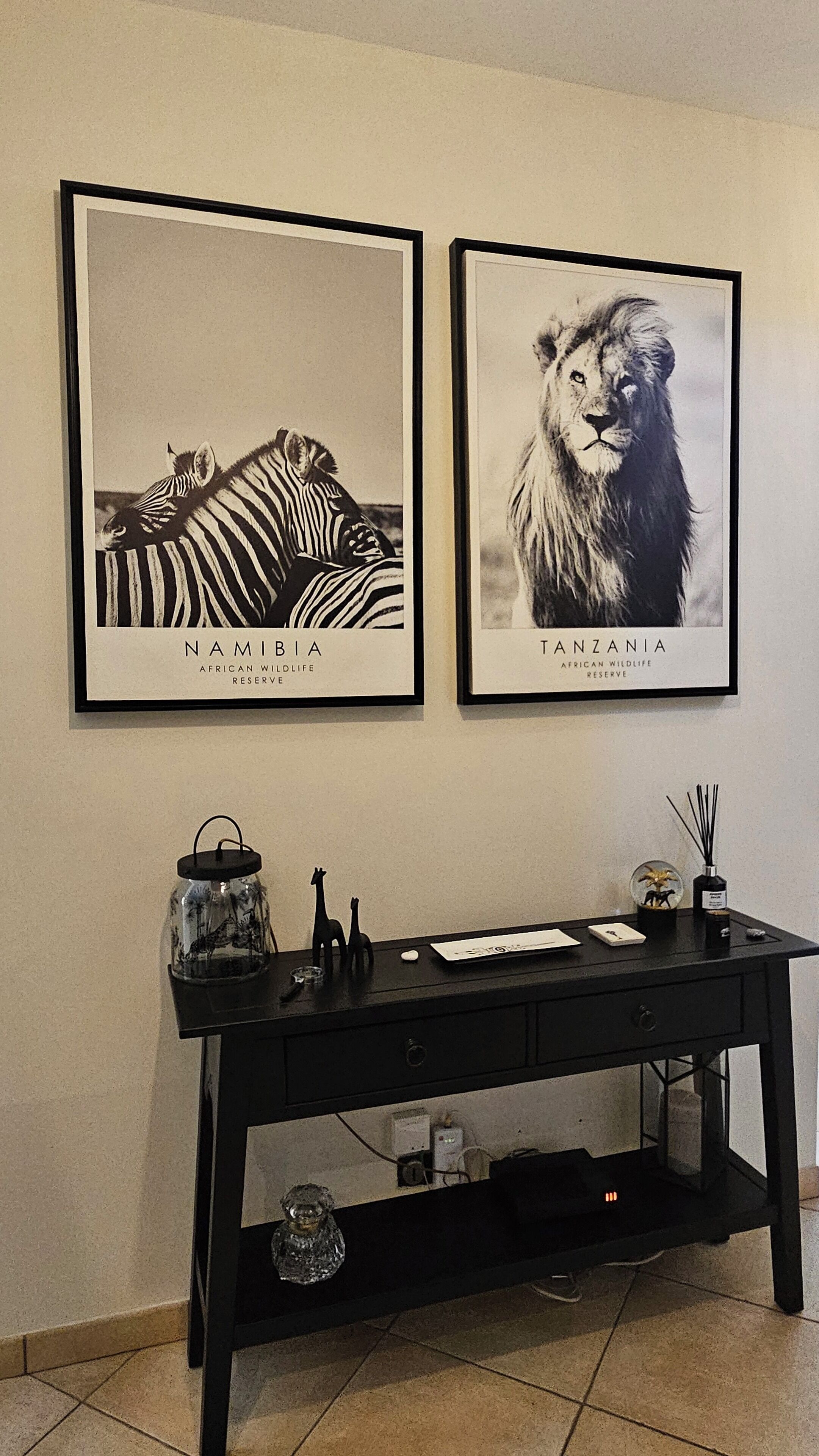 Two framed wildlife photographs labeled 'Namibia' and 'Tanzania' above a desk with a lamp, a glass container with figurines, and a small plant.