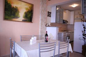 Uma área de refeições com uma mesa posta para dois, um quadro na parede e uma cozinha ao fundo.