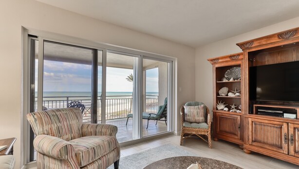 Een woonkamer met een grote schuifpui naar een balkon met uitzicht op de oceaan. Er staat een gestreept fauteuil, een rieten stoel en een houten tv-meubel met een flatscreen televisie.