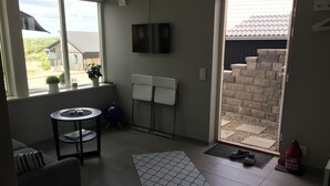 A living room with a sofa, a small round table, a TV mounted on the wall, and a sliding glass door leading to an outdoor area with a brick wall and a garden. - Functional apartment with sea view (Tanum)