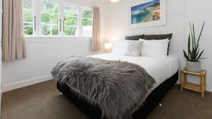 A bedroom with a large bed, a window with curtains, a wall-mounted lamp, a framed picture, and a small plant on a side table. - Private Bay Beachfront - Stunning Location! (Coromandel Township)
