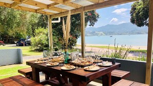 A wooden deck with a table set for dining, overlooking a lake and mountains. - Private Bay Beachfront - Stunning Location! (Coromandel Township)