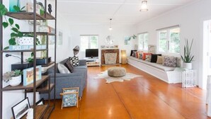 A spacious living room with a grey sofa, wooden flooring, and a bookshelf filled with plants and books. - Private Bay Beachfront - Stunning Location! (Coromandel Township)