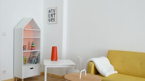 Ein Zimmer mit einem weißen Bücherregal, einem kleinen weißen Tisch, einem gelben Sofa und einem Schild mit der Aufschrift 'ADVENTURE'.