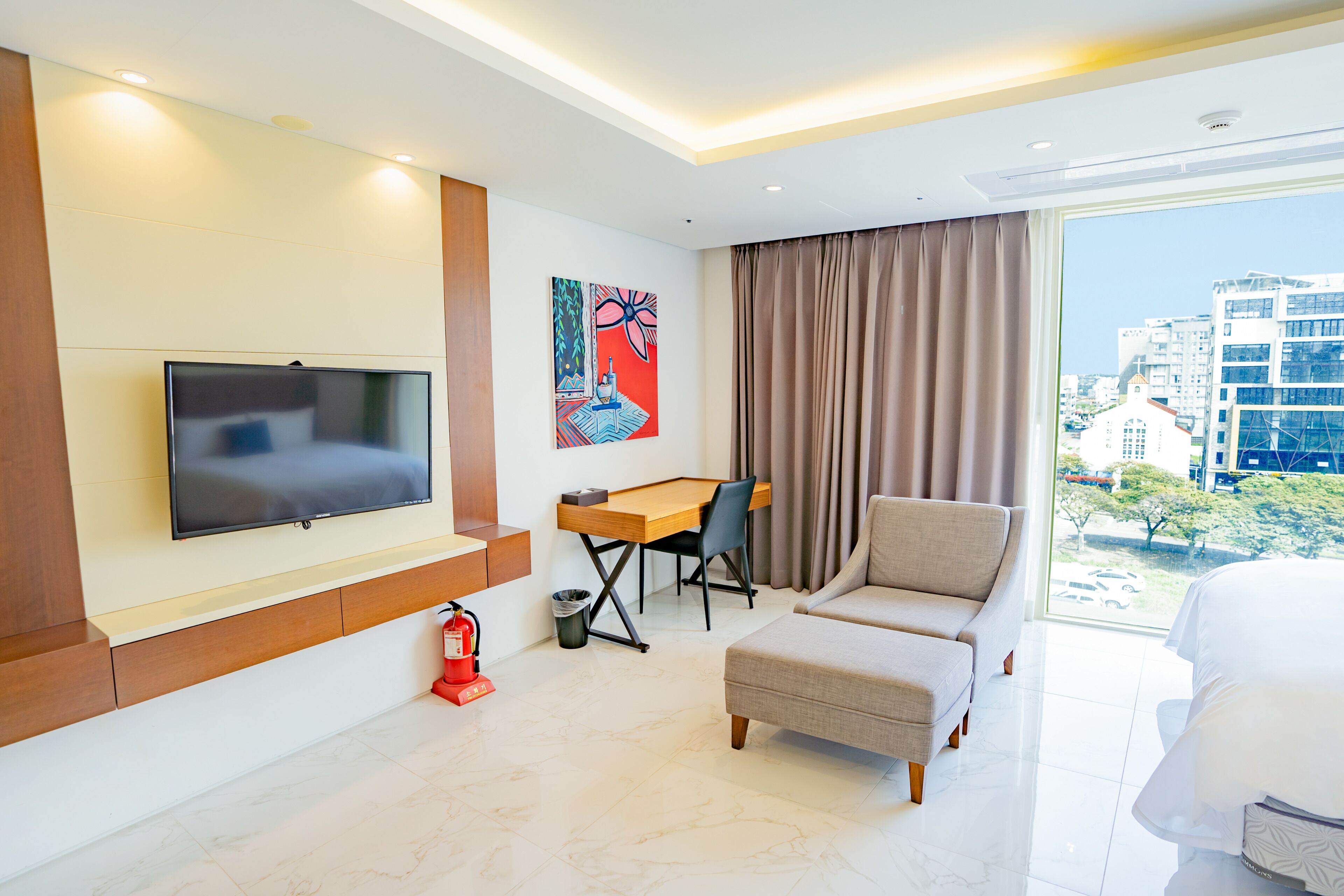 A modern hotel room with a flat-screen TV mounted on the wall, a wooden desk with a chair, a beige armchair, and a large window offering a city view.