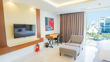 A modern hotel room with a flat-screen TV mounted on the wall, a wooden desk with a chair, a beige armchair, and a large window offering a city view.