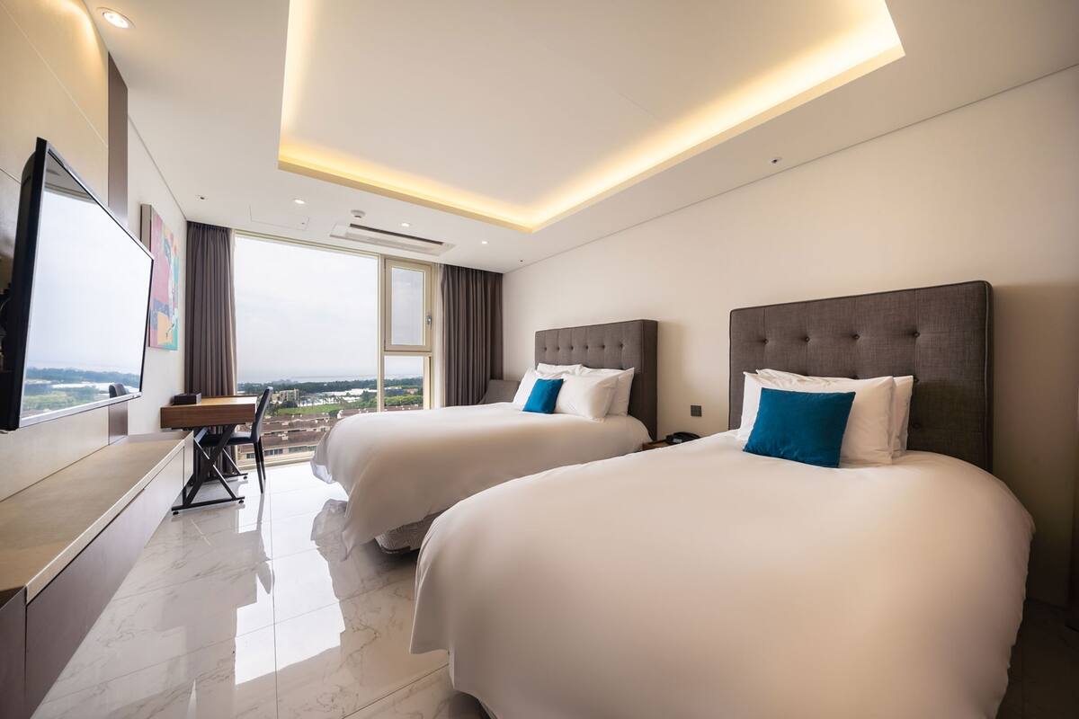 Habitación de hotel con dos camas, un escritorio y un ventanal grande que ofrece vistas a la ciudad.