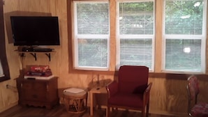 Una habitación con un televisor montado en la pared, un sillón rojo, una mesita redonda y una silla de mimbre.
