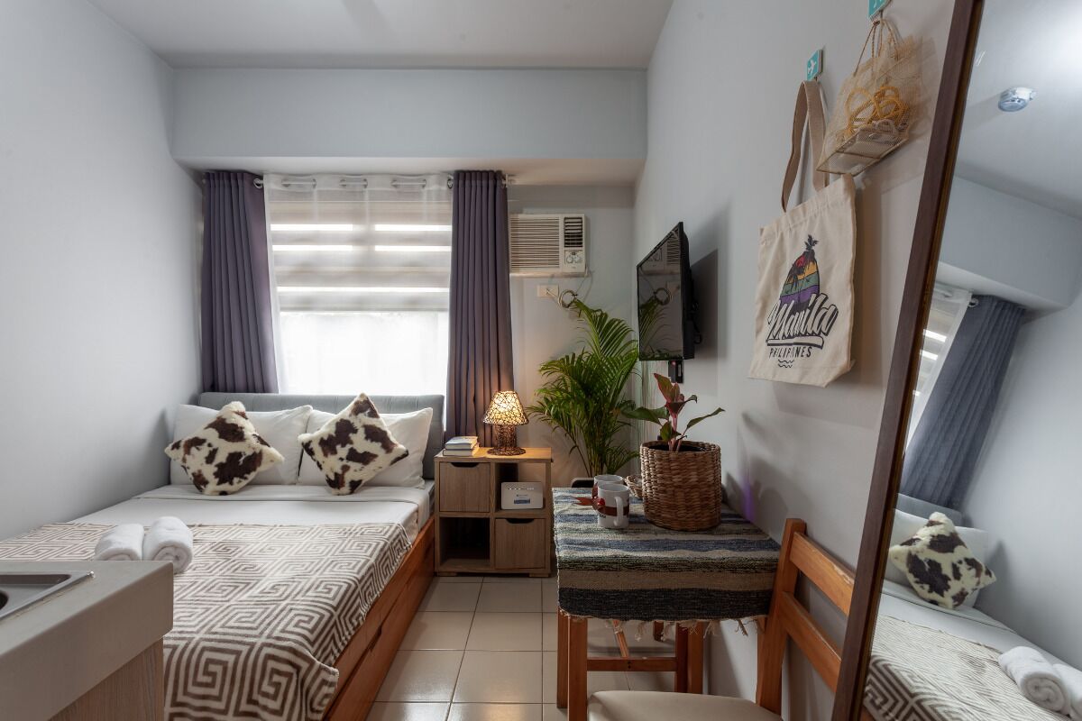 A compact hotel room with a bed, a small table, a chair, a potted plant, a wall-mounted TV, and a window with curtains.