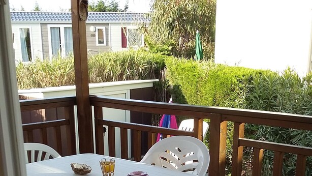 Een overdekt terras met een witte tafel en stoelen, omgeven door een houten terras en een uitzicht op groen en gebouwen in de verte.