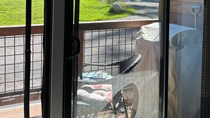 A view through a glass door of a patio area with a grill, outdoor furniture, and a grassy landscape.
