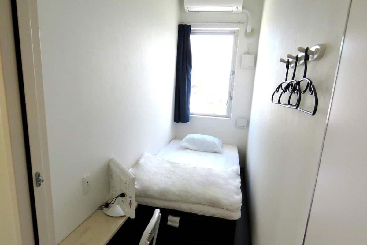 A small, narrow room with a single bed, a bedside table with a lamp, a window with a curtain, and a coat rack with hangers.