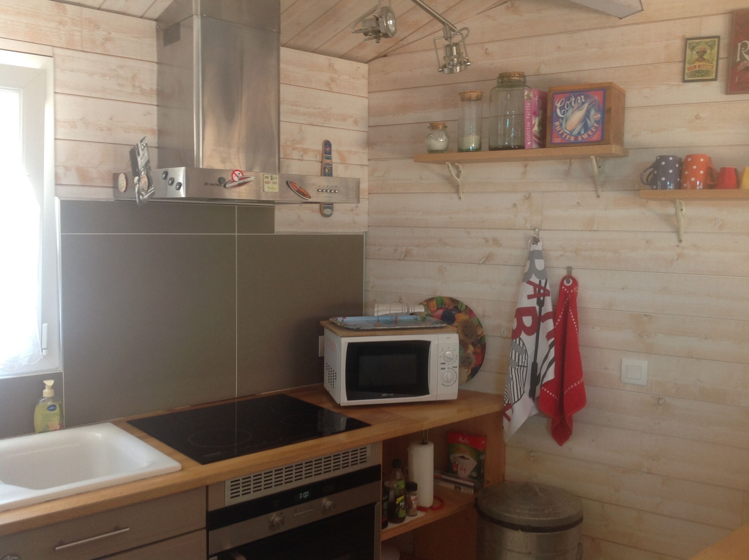 A kitchen with wooden walls, a stainless steel range hood, a microwave, and various kitchen utensils on shelves.