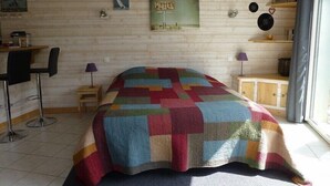 A cozy room with a wooden wall, a colorful quilted bedspread, a small table with a lamp, and a wall clock.