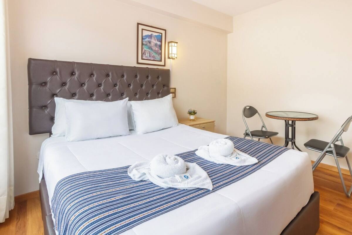 Ein Hotelzimmer mit einem großen Bett, einem Kopfteil, einem Nachttisch mit Lampe, einem kleinen runden Tisch mit Stühlen und einem gerahmten Bild an der Wand.
