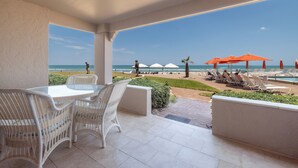 Un espacio al aire libre techado con una mesa y sillas, con vista a una playa con sombrillas y sillas de descanso.