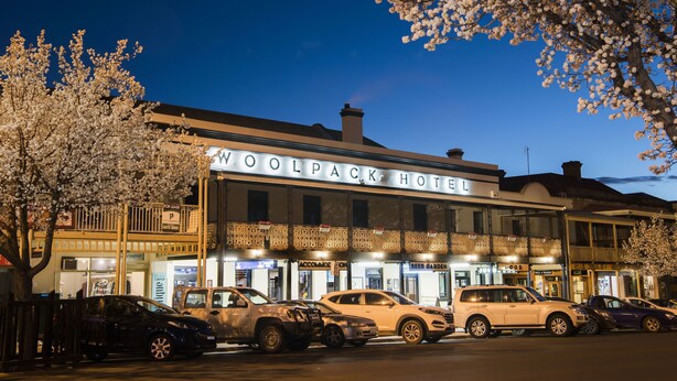 The Woolpack Hotel with a parking lot in front, a sign for a nearby restaurant, and a tree with blossoms to the left.