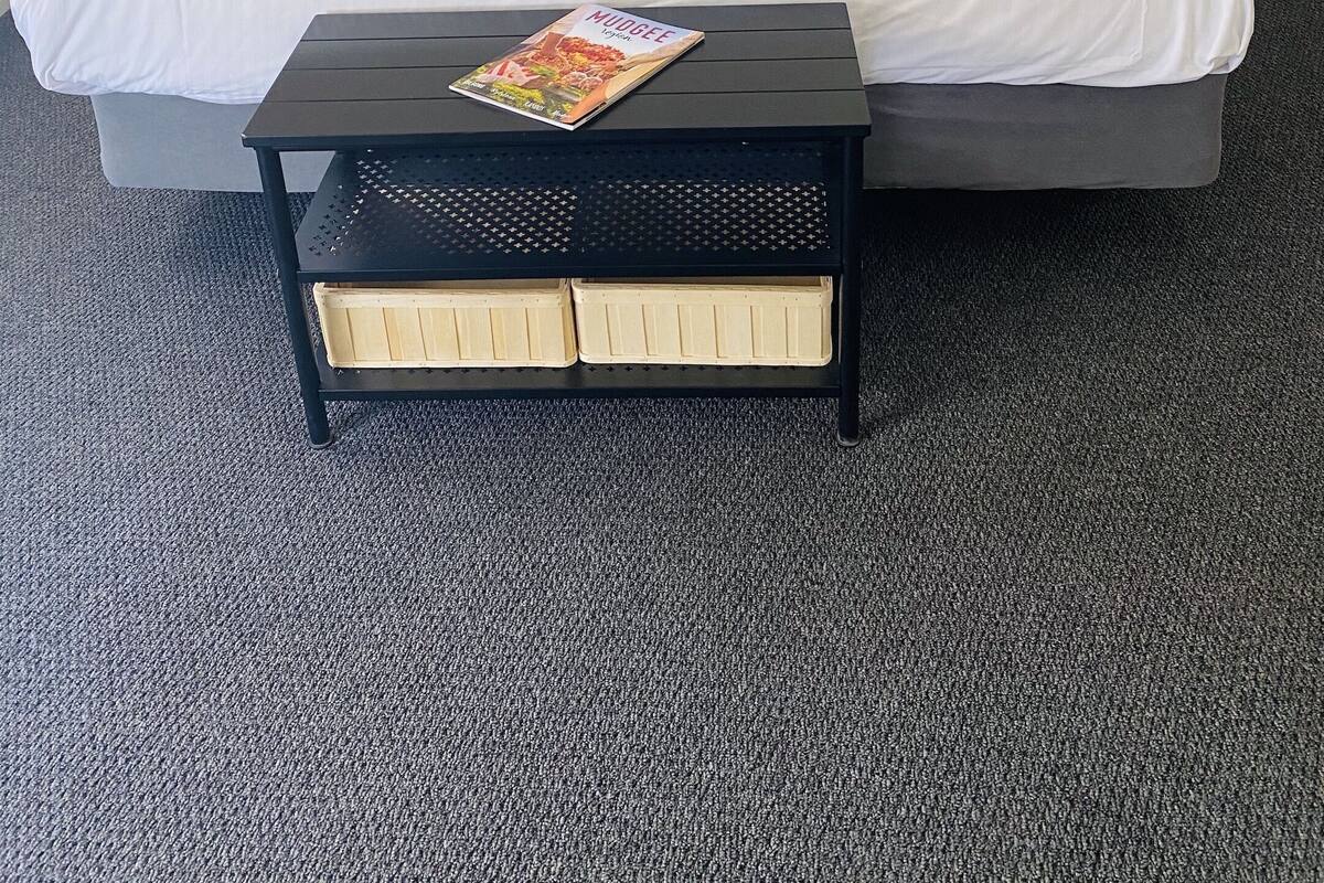 A neatly made bed with two pillows, a dark throw blanket, and a magazine on a small table beside it.