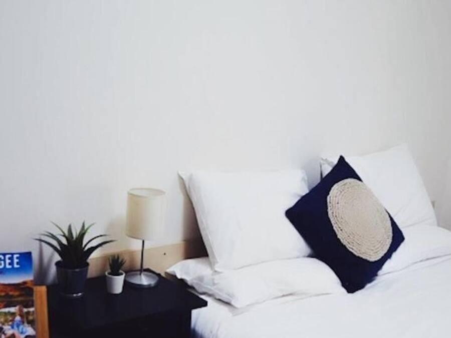 A neatly made bed with white linens and a dark pillow, a small bedside table with a lamp, a potted plant, and a book titled "Mudgee."