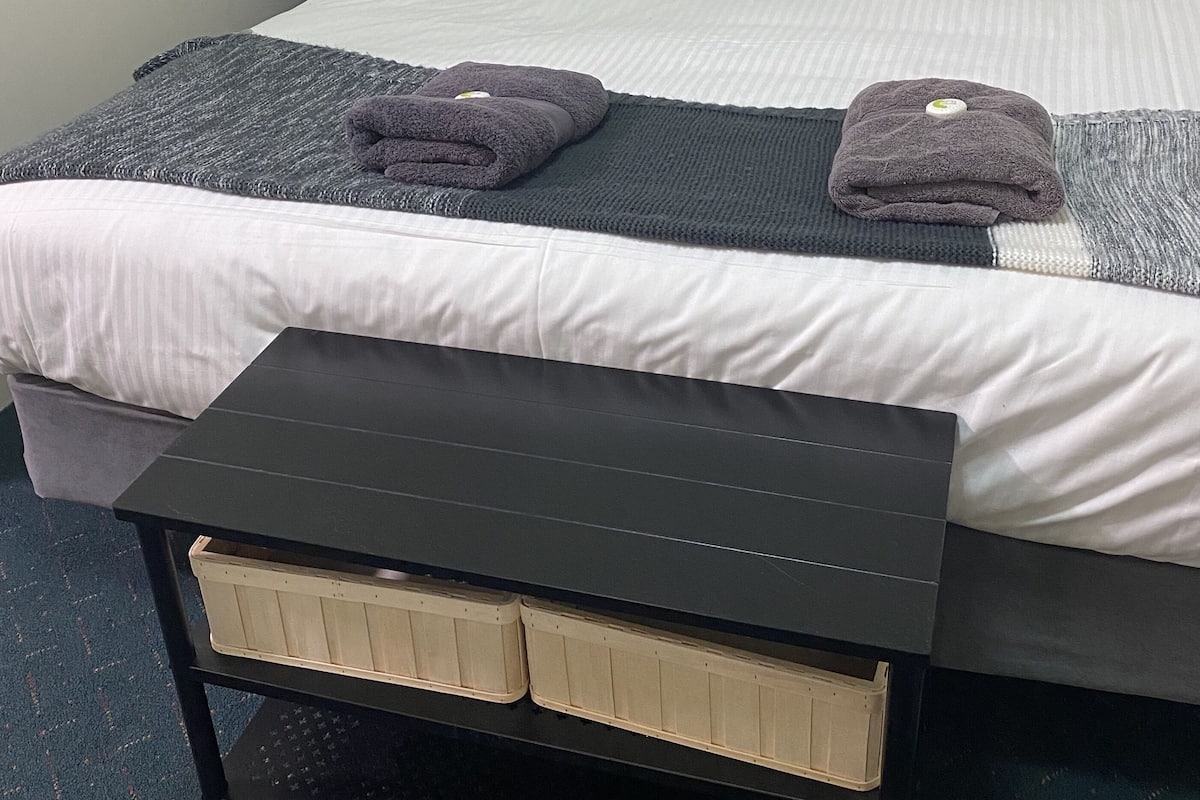 A neatly made bed with white and gray bedding, a blue pillow, and two folded towels. There is a small bedside table with a lamp and a dark-colored bench with storage drawers.