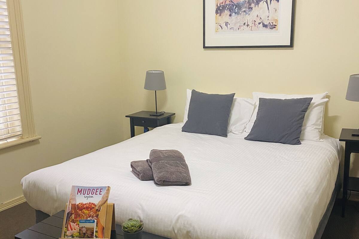 A neatly made bed with pillows, a framed picture on the wall, and a book on the nightstand.