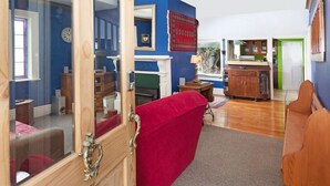 A room with a red sofa, a wooden door, a mirror, a clock, and a kitchen area in the background.