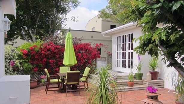 A patio with a brick floor, a table with chairs, and a green umbrella. There are potted plants and a white building in the background. - Fab Studio Plus!!- Pukekura Park/CBD right there (New Plymouth)