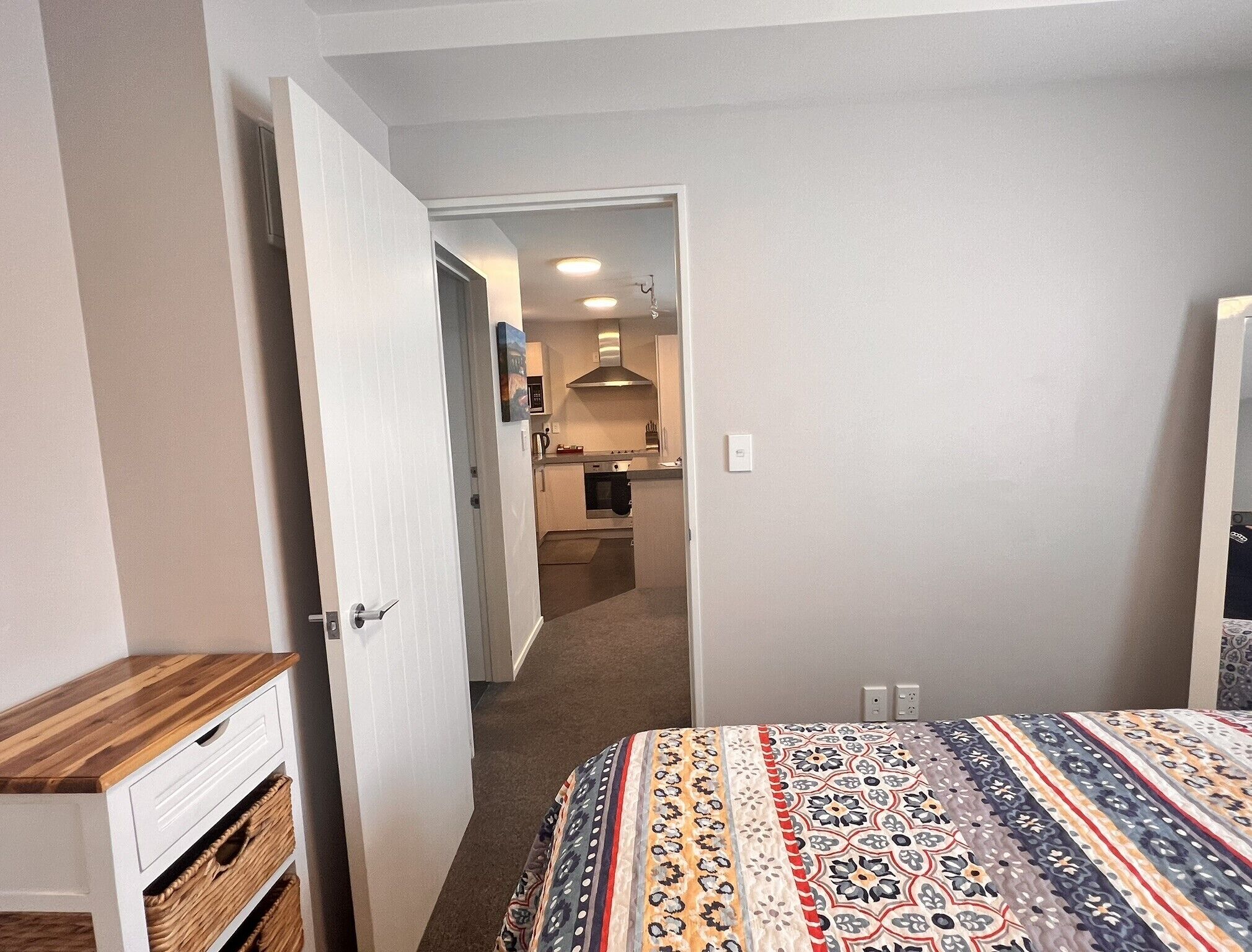 Une chambre d’hôtel avec un lit, une table de chevet en bois, une porte donnant sur une salle de bain et, au fond, une petite cuisine.