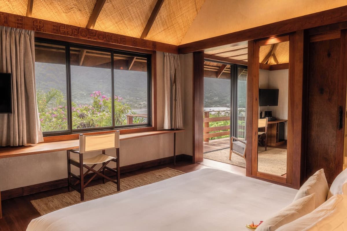 A bedroom with a large bed, a desk, a chair, a TV, and a balcony with a view.
