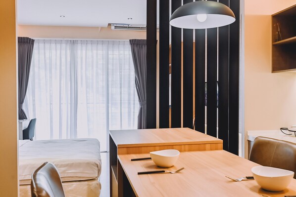 Habitación de hotel con una cama, una mesa de comedor con palillos y cuencos, una lámpara colgante negra y una ventana con cortinas.