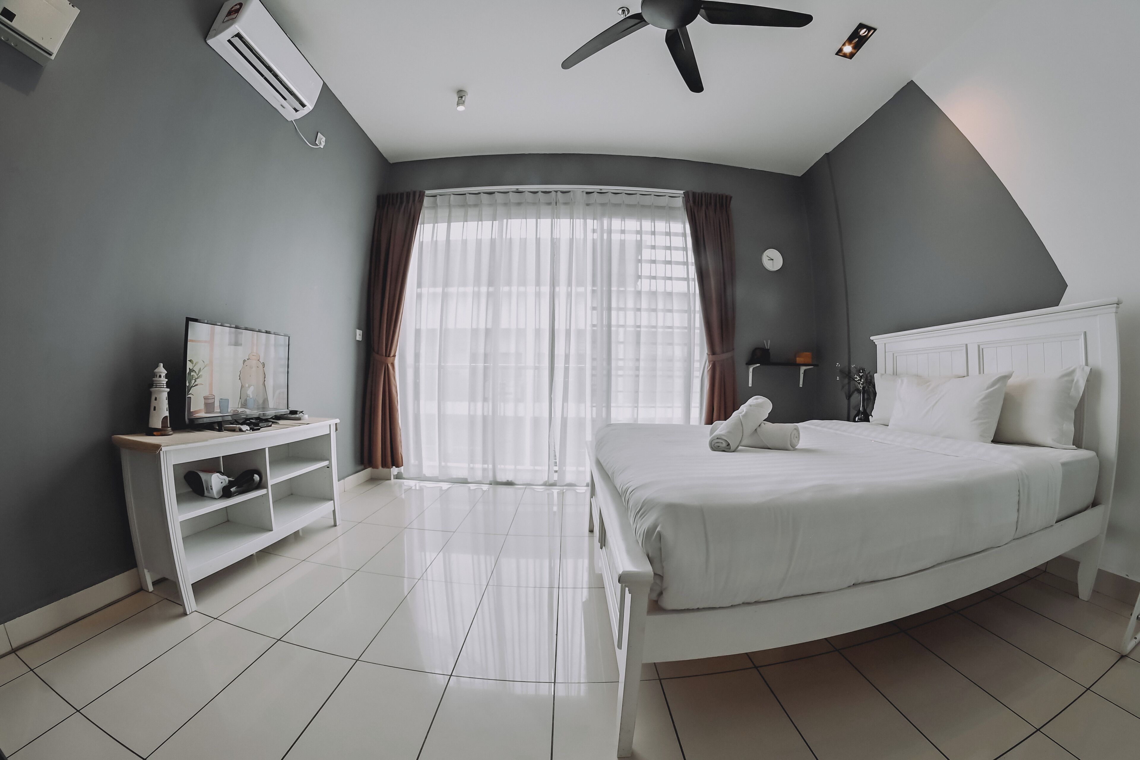 Una habitación de hotel moderna con una cama curva, un televisor sobre un mueble blanco, un ventilador de techo y un ventanal grande con cortinas.