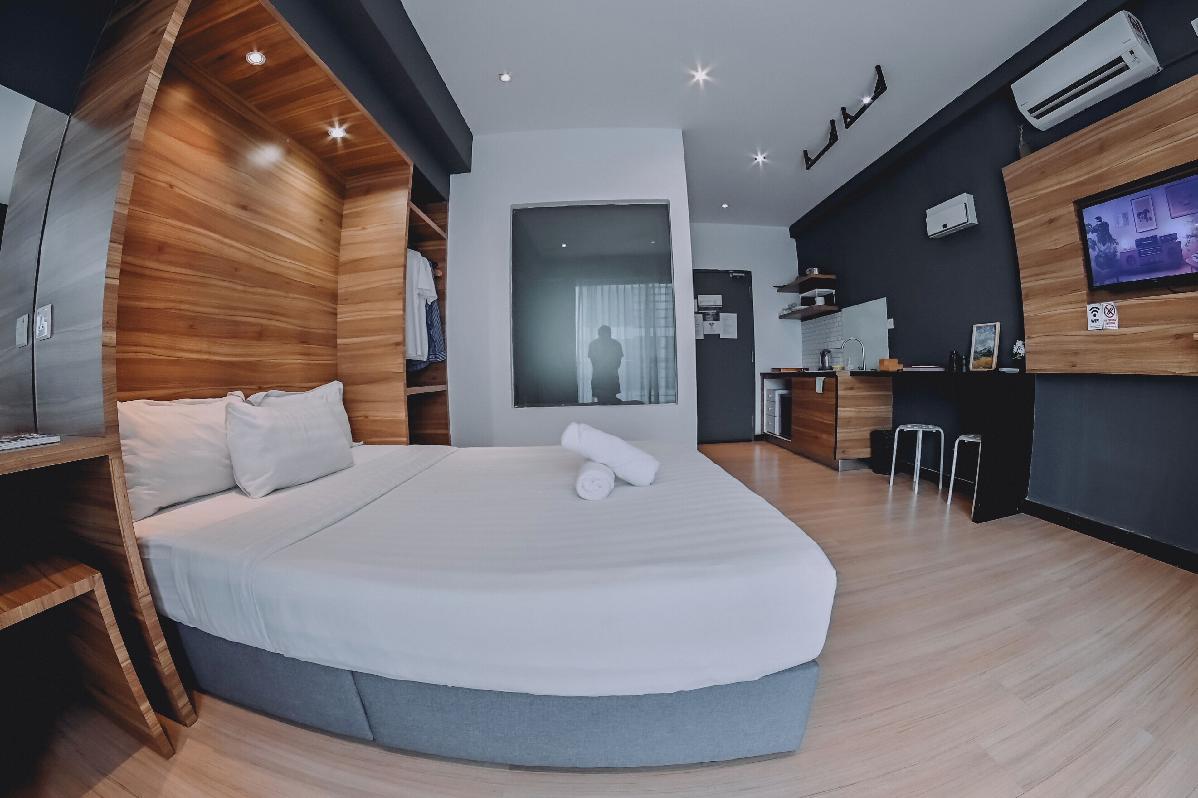 A modern hotel room with a curved bed, wooden paneling, a flat-screen TV, a small kitchenette, and a large window.