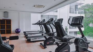 Gimnasio con cintas de correr, elípticas y una máquina de remo. Hay una pelota de ejercicios morada y un armario de madera. La sala tiene ventanas amplias con vistas a edificios y zonas verdes.